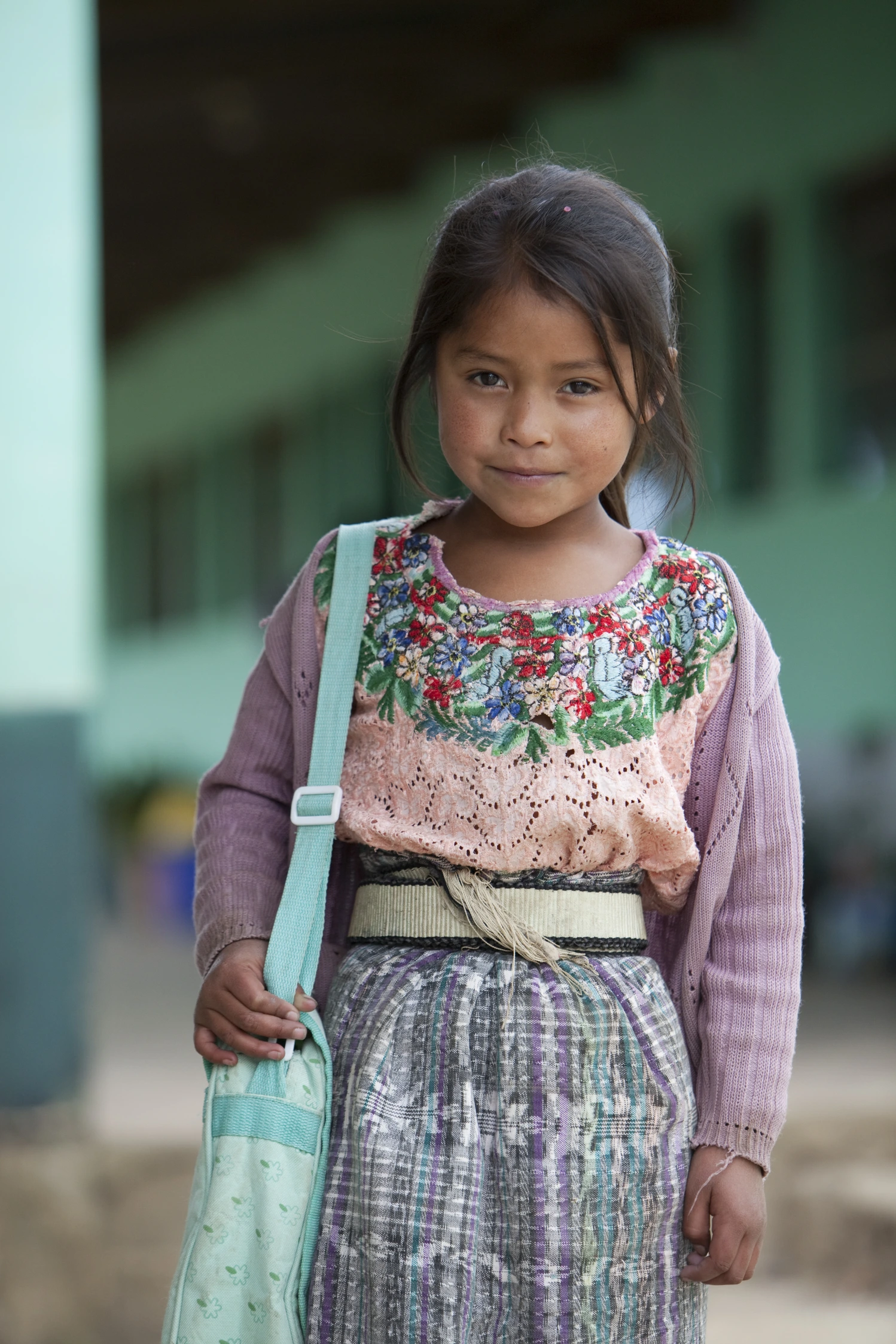 A young girl, wearing a traditional embroidered outfit and carrying a light blue bag, smiles softly while standing outside.