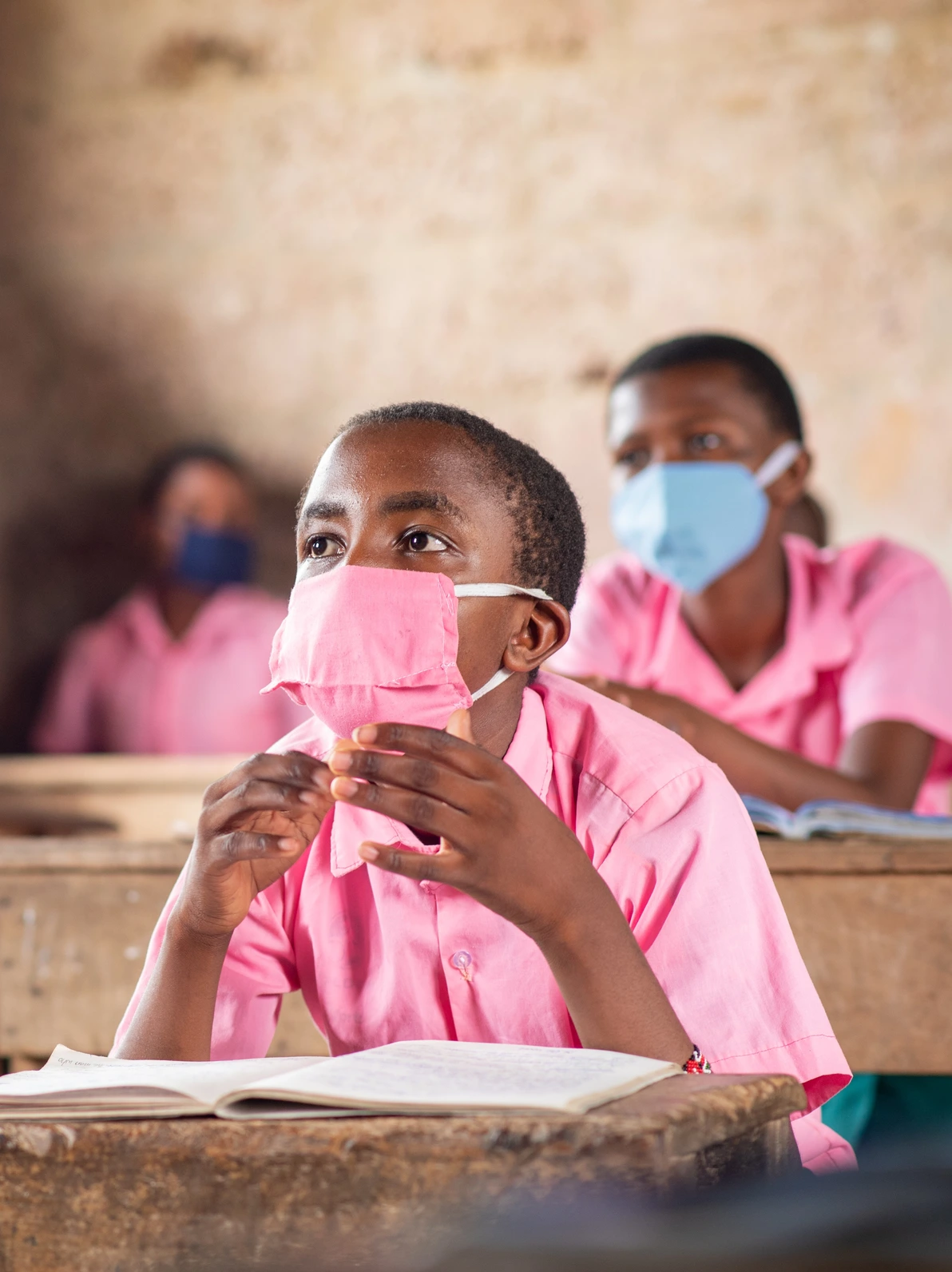 A student wearing a face mask and a pink uniform sits attentively in class with a book open in front of them.