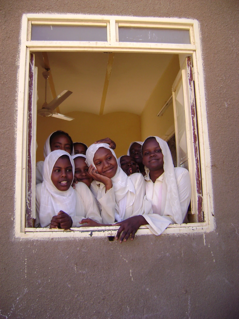 A group of smiling young girls wearing white hijabs look out from a window in a light-colored building.