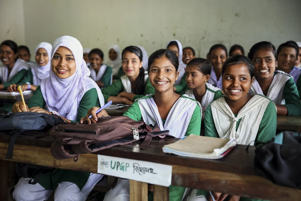 A classroom full of smiling young female students in green and white uniforms, with some wearing hijabs, sitting at desks with school supplies.